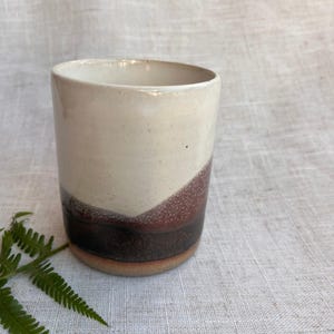 Peut inclure: Un gobelet en céramique fait à la main avec un dégradé de couleurs unique. La moitié supérieure est blanc crème, passant à un marron moucheté puis à une base marron foncé. Le gobelet a une forme lisse et arrondie.