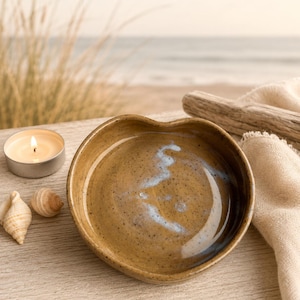 May include: A heart-shaped ceramic dish with a speckled tan glaze and a subtle blue swirl pattern in the center. The bowl has a smooth, glossy finish and a slightly irregular rim, reflecting a handcrafted appearance with earthy tones.