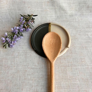 May include: A wooden spoon rests on a two-toned ceramic spoon rest. The spoon rest is divided into black and cream sections with a gold accent. A sprig of purple rosemary adds a natural touch to the composition.