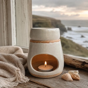 May include: A cream-colored ceramic wax warmer with a glossy finish and a wide, arched opening at the base. The warmer features a top dish for wax melts and a lit tea light candle inside the central cavity, showcasing a rustic, unglazed rim.