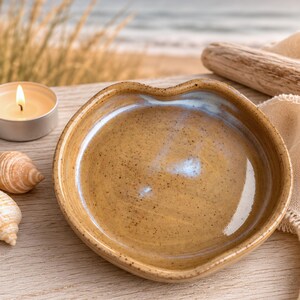 May include: A round, shallow ceramic dish with a wavy, organic rim. The glaze is a warm, speckled tan color with subtle blue-toned highlights in the center. The surface has a glossy finish, and the clay body shows fine dark speckles throughout.