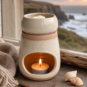 May include: A cream-colored ceramic wax warmer with a smooth glazed finish and an exposed terracotta rim. The base features a wide opening containing a lit tea light candle, while the top bowl holds two white wax melts for fragrance diffusion.