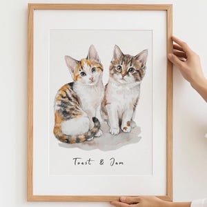 Può includere: Un'illustrazione ad acquerello incorniciata di due gattini seduti fianco a fianco. Il gattino a sinistra è tricolore con pelo arancione, nero e bianco, mentre quello a destra è un tabby marrone. Il testo "Toast & Jam" è stampato in nero sotto i gatti.