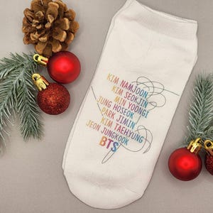 Peut inclure: Chaussette blanche avec les noms des membres de BTS en arc-en-ciel. La chaussette est ornée d'un dessin au trait et entourée de décorations de Noël et d'aiguilles de pin.