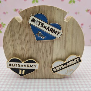 Op de afbeelding: Drie hartvormige emaille pins met de tekst "#BTSxARMY". Eén pin is blauw met de tekst "RM", een andere is zwart en de derde is wit met de naam "Jin". De pins worden getoond op een houten standaard.