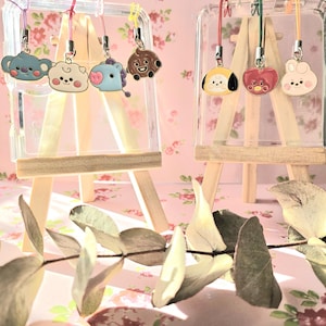 Könnte beinhalten: Eine Präsentation von bunten Cartoon-Charakter-Anhängern, die an transparenten Acrylstaffeleien hängen. Die Anhänger zeigen verschiedene Tierdesigns in Blau-, Weiß-, Braun-, Gelb- und Rottönen. Der Hintergrund ist ein rosa Blumenmuster.