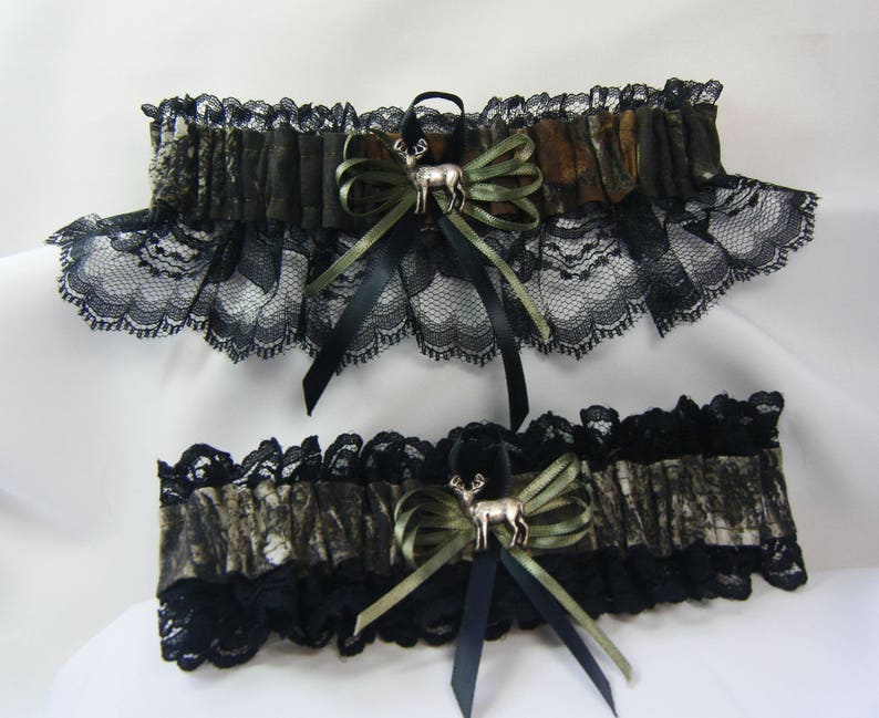 May include: Two-piece set featuring a camouflage pattern with black lace trim. Each piece has a decorative bow with a silver deer charm. The bows are accented with olive green and black ribbons. The set is designed with a rustic theme.