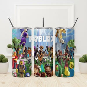 May include: Three stainless steel tumblers with a Roblox theme. Each tumbler features a different design with Roblox characters and the Roblox logo. The tumblers have a silver straw and a lid. The background shows a city scene.