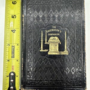 May include: A black leather-bound book with gold lettering and a gold embossed image of a Masonic altar with candles and a compass and square. The title of the book is "The Freemason's Companion."