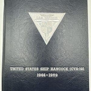 May include: A black leather-bound book with a silver triangle emblem that reads "USS. HANCOCK" and a silver crest. The book also has the text "UNITED STATES SHIP HANCOCK (CVA-19) 1944-1969" printed on the front cover.