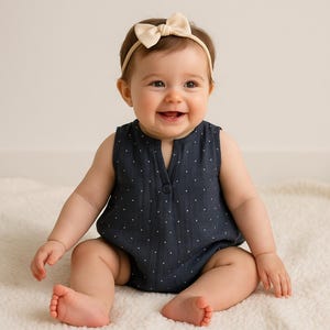 May include: A baby wearing a navy blue sleeveless romper with white polka dots and a cream-coloured bow headband. The romper has a v-neck and a single button closure. The baby is sitting on a white, fluffy surface.
