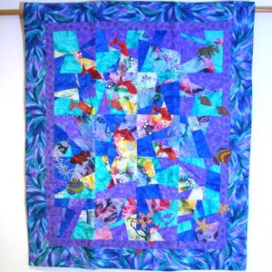 Puede incluir: Una colcha colorida con un patrón geométrico de cuadrados y triángulos azules, morados y turquesa. La colcha presenta una variedad de diseños florales y de peces.