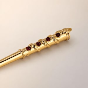 May include: A gold-toned metal hair stick featuring a coiled snake design wrapped around the upper portion. The snake body is textured with scales and is accented with four round, deep red cabochon stones set along its length.