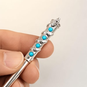 May include: A silver-toned metal hair stick featuring a coiled snake design wrapped around the shaft. The snake is accented with four round, bright turquoise-colored cabochon stones set in a vertical row along the length of the hair accessory.