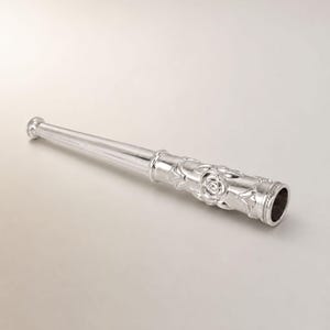 May include: A silver-coloured cigarette holder with a tapered, smooth stem and a cylindrical end featuring a raised, embossed rose floral pattern and decorative leaf motifs. The object has a polished, metallic finish and a hollow opening at the end.