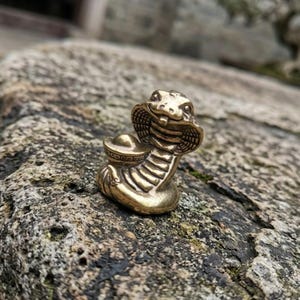 May include: A small brass figurine of a cobra snake with its hood flared, holding a traditional gold ingot in its coils. The metal has a polished, warm golden finish with detailed etched lines along the body and hood of the snake sculpture.