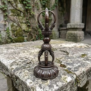 Puede incluir: Un objeto ritual vajra de metal color bronce oscuro que presenta un mango central con intrincadas tallas en relieve, rematado con una corona de múltiples puntas y apoyado sobre una base circular en forma de pétalo con grabados detallados.