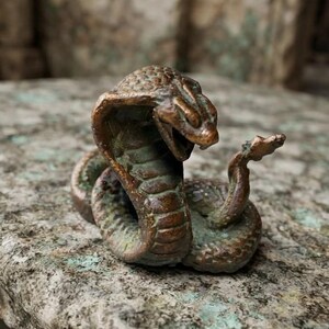 Könnte beinhalten: Eine kleine, zusammengerollte kobraförmige Figur in Bronzefarbe mit erhobener Haube und offenem Maul. Die Metalloberfläche weist eine verwitterte, dunkelbraune und grüne Patina mit sichtbaren Schuppentexturen am Körper und einem aufwärts gebogenen Schwanz auf.