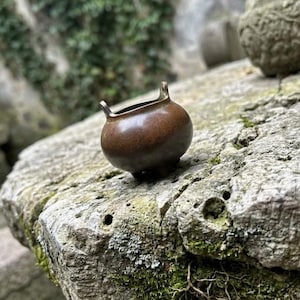 以下が含まれることがあります： 滑らかで光沢のある表面を持つ、ブロンズ色の小さな丸い金属製の容器。上部の縁には2つの小さな垂直なハンドルがあり、3本の短く頑丈な脚で支えられており、コンパクトでバランスの取れたシルエットを形成しています。