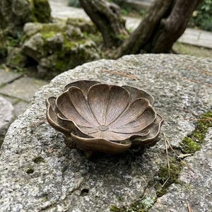 May include: A bronze-toned metal bowl shaped like a blooming flower with layered, textured petals. The interior features fine, radiating lines that converge at a central point, and the exterior base is supported by small, rounded feet.
