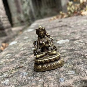 以下が含まれることがあります： 瞑想のポーズで座っている神の小さな真鍮色の金属製フィギュア。王冠、装飾品、衣服に複雑なディテールが施されており、蓮の形をした台座の上で両手を祈りのポーズで組んでいる姿が表現されています。
