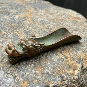 Pode incluir: Uma colher de metal em forma de dragão com uma pátina de bronze e zinabre envelhecida. O objeto apresenta uma cabeça de dragão detalhada numa extremidade com chifres e focinho, que se estende para um cabo curvo e aberto para servir.