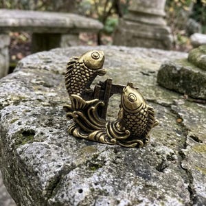 以下が含まれることがあります： 真鍮色の金属製フィギュアで、彫刻された波から飛び出す2匹の様式化された魚が表現されています。魚は互いに向き合い、詳細な鱗とひれが施されており、中央の小さな垂直構造によって連結された一体感のあるデザインとなっています。