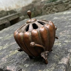 May include: A bronze-toned metal incense burner shaped like a pumpkin with vertical ridges. The lid features a central knob and cut-out openings. The base is supported by three legs sculpted to resemble plant stems with small leaves and buds.