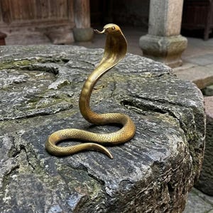 Puede incluir: Una figura de cobra de latón en postura erguida y alerta con la capucha desplegada. La serpiente presenta texturas de escamas detalladas a lo largo del cuerpo, una cabeza lisa y una lengua bífida que sale de la boca, en tono dorado.