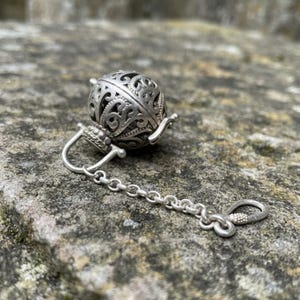 May include: A silver-toned spherical charm featuring an intricate openwork filigree pattern. The charm is attached to a short, delicate metal chain that ends in a small, oval-shaped clasp, creating a cohesive piece of jewelry hardware.