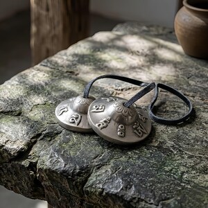 May include: Two silver-colored Tibetan Tingsha cymbals with embossed symbols and a black leather cord. The cymbals are resting on a textured, gray stone surface. The cymbals are used in meditation and sound healing.