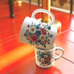 May include: Two white ceramic mugs with floral designs. The mugs feature a bouquet of colorful flowers in shades of red, blue, and yellow. The mugs have a curved handle and are set against a red surface.