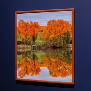 May include: A framed landscape photograph featuring a serene lake reflecting vibrant autumn foliage. The trees display shades of orange and yellow, mirrored in the water. A small dock is visible, set against a backdrop of green hills and a cloudy sky.