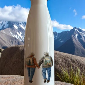 May include: White insulated water bottle with a silver lid. The bottle features a sticker of two people holding hands, wearing backpacks with a mountain scene. The background shows a mountain range and blue sky.