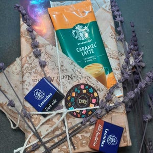 Può includere: Un pacchetto regalo avvolto in carta con motivo a mappa contenente una bustina di caffè istantaneo Starbucks Caramel Latte, due quadratini di cioccolato Leonidas etichettati Dark Nibs e Milk Feuilletine, un adesivo circolare Love e un segnalibro.