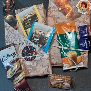 May include: A gift package wrapped in map-patterned paper featuring Twinings Creamy Vanilla and Vanilla Chai tea bags, a Starbucks Caramel Latte packet, a Galaxy Instant Hot Chocolate sachet, two Leonidas chocolates, and a BeBe Petit Beurre biscuit.