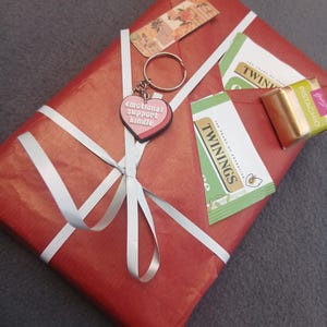 May include: A rectangular gift wrapped in red paper with a white ribbon, featuring a heart-shaped keychain with the text emotional support kindle, two Twinings tea bags, a small floral tag, and a gold-wrapped pistachio chocolate square.