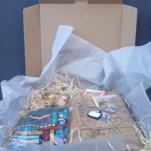 May include: A cardboard gift box containing a folded dark fabric item with a colorful geometric pattern, a rectangular package wrapped in brown paper with vintage travel-themed prints, Twinings tea bags, and a small black and white keychain.