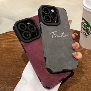 Peut inclure: Deux coques de téléphone avec une finition en daim texturé. L'une est grise avec le nom "Freda" écrit en blanc. L'autre est bordeaux. Les deux coques ont un cache objectif d'appareil photo noir avec plusieurs objectifs.