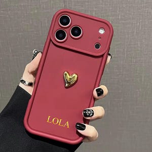 Peut inclure: Coque de téléphone bordeaux avec un cœur doré et le nom "LOLA" en lettres dorées. L'étui a un bord surélevé autour des objectifs de l'appareil photo et un cadre protecteur.