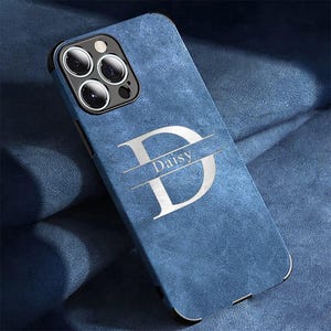 Puede incluir: Funda de teléfono azul con un acabado texturizado. La funda presenta una gran letra plateada "D" con el nombre "Daisy" en el centro. La funda tiene un borde negro y recortes para la cámara del teléfono.