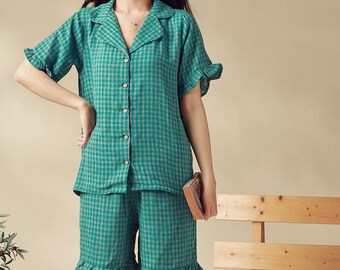 May include: A woman wearing a green and white checkered pajama set with short sleeves and shorts. The shorts have a ruffle trim. She is standing in front of a wooden bench.