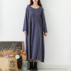 May include: A long, slate blue dress with long sleeves and a round neckline. The dress features vertical pleats down the front and a loose, flowing silhouette. The model is wearing black ankle boots.