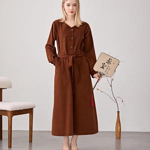 May include: A long-sleeved, brown dress with a V-neck, button closure, and a matching belt. The dress has pockets and is worn with beige flats. A decorative fan with Chinese characters is held in the hand.