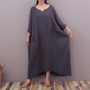 May include: A dark gray, long-sleeved dress with a V-neck and two large front pockets. The dress is ankle-length and appears to be made of a soft, flowing fabric. The sleeves are rolled up at the elbows. The person is wearing brown shoes.
