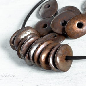 May include: A close-up of a necklace made with a stack of oval-shaped, copper-toned ceramic beads. The beads have a shiny, metallic finish and are strung on a black cord.