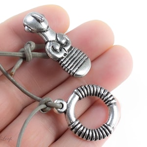 Puede incluir: Un collar de plata con dos colgantes. Un colgante es una figura humana estilizada y el otro es un círculo con un diseño texturizado. El collar está hecho de un cordón de cuero gris.