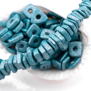 May include: A close-up of teal blue, square-shaped beads with a metallic sheen. The beads are strung together to form a necklace or bracelet. The beads are scattered around a white seashell.
