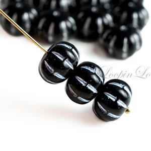 Op de afbeelding: Zwarte glazen kralen met witte strepen. De kralen zijn rond en hebben een getextureerd oppervlak. De kralen zijn geregen aan een gouden draad.