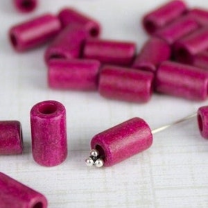 Puede incluir: Primer plano de un grupo de cuentas de madera de color rosa. Las cuentas son cil&iacute;ndricas y tienen un acabado liso y pulido. Algunas de las cuentas tienen una peque&ntilde;a bola de metal en su interior.